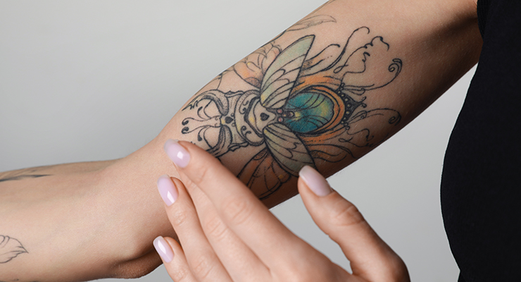 Woman applying cream on her arm with tattoos against light background, closeup